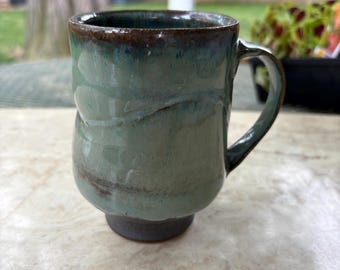 Op de afbeelding: Handgemaakte keramische mok met een turquoise en bruin glazuur. De mok heeft een uniek, getextureerd ontwerp en een stevig handvat. De basis is een donkerdere blauwgrijze tint. Perfect voor koffie of thee.