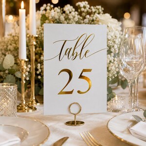 May include: A white rectangular table number card featuring the gold foil text Table 25 in a script and serif font combination. The card is held upright by a small gold circular metal stand, positioned on a table setting with gold accents.