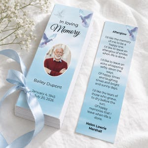 May include: A funeral programme bookmark with a light blue sky background, white doves, and the text In loving Memory. It features a circular photo of a person, the name Bailey Dupont, dates, and the poem Afterglow by Helen Lowrie Marshall.