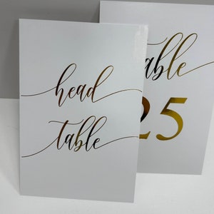 May include: Two white table cards with gold lettering. The first card says "head table" in cursive. The second card says "table 25" in cursive.