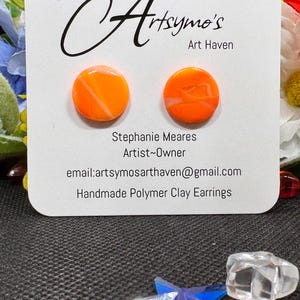 May include: Two round, bright orange stud earrings with subtle white marbled patterns, displayed on a white card labeled Artsymo's Art Haven, Stephanie Meares Artist~Owner, email:artsymosarthaven@gmail.com, and Handmade Polymer Clay Earrings.