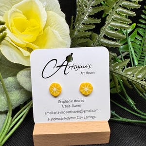 May include: A pair of small, round, yellow lemon slice earrings made of polymer clay, displayed on a white card labeled Artsymo's Art Haven by Stephanie Meares, featuring the email artsymosarthaven@gmail.com and the text Handmade Polymer Clay Earrings.