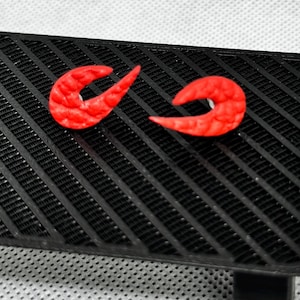 May include: Two bright red, curved earrings with a textured surface rest on a black, slatted surface. The earrings have a unique, abstract shape. The black surface has a grid-like pattern.