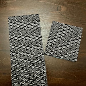 Pode incluir: Dois artigos retangulares de tecido com um padrão de ondas a preto e branco. A peça maior está em pé, e a peça menor está angulada para a direita. O padrão consiste em arcos brancos repetidos sobre um fundo preto.