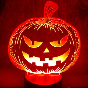 May include: A red, orange, and yellow 3D printed pumpkin shaped night light with a carved jack-o-lantern face. The light is on a black base with a button on the side. A remote control with multiple buttons is in the foreground.