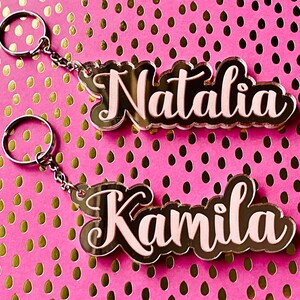May include: Two acrylic keychains with the names "Natalia" and "Kamila" written in black with a pink outline. The keychains are on a pink background with gold polka dots.