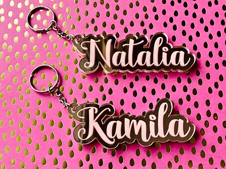 May include: Two acrylic keychains with the names "Natalia" and "Kamila" written in black with a pink outline. The keychains are on a pink background with gold polka dots.