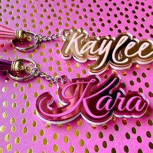May include: Two pink and white acrylic keychains with the names "Kaylee" and "Kara" written in cursive. Each keychain has a silver chain and a tassel.
