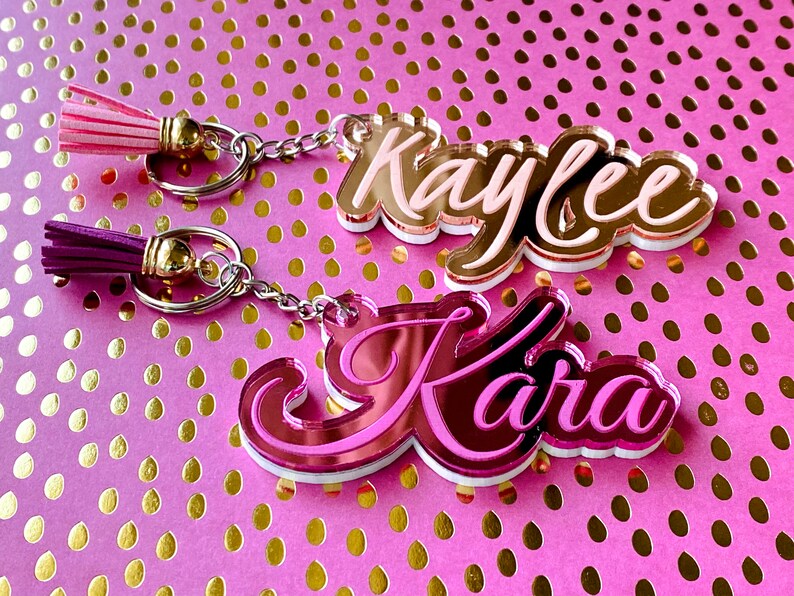 May include: Two pink and white acrylic keychains with the names "Kaylee" and "Kara" written in cursive. Each keychain has a silver chain and a tassel.