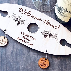 May include: A wooden wine bottle holder with a welcome home message, grapevine design, and a realtor's contact information. It includes two wooden tags that say "Welcome Home!" and "Home Sweet Home".