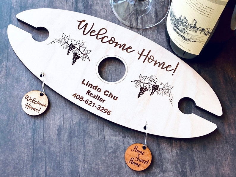 May include: A wooden wine bottle holder with a welcome home message, grapevine design, and a realtor's contact information. It includes two wooden tags that say "Welcome Home!" and "Home Sweet Home".