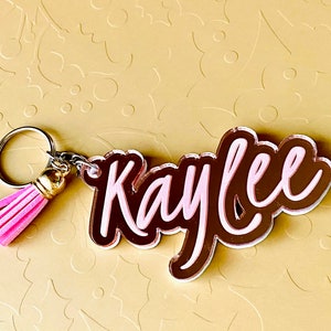 May include: A pink and brown acrylic keychain with the name "Kaylee" in a cursive font. The keychain has a pink tassel attached to a silver key ring.