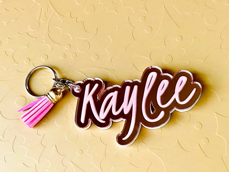 May include: A pink and brown acrylic keychain with the name "Kaylee" in a cursive font. The keychain has a pink tassel attached to a silver key ring.