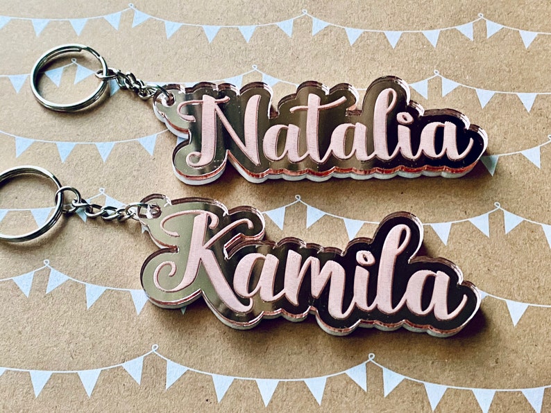 May include: Two silver keychains with rose gold mirror acrylic charms. The charms are shaped like the names "Natalia" and "Kamila" in a cursive font. The names are filled with a light pink color.