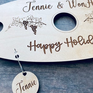 May include: Personalized wooden serving tray with two wine glass holders and a grapevine design. The tray says "Jennie & Won" and "Happy Holidays". A small wooden tag with the name "Jennie" is attached to the tray.