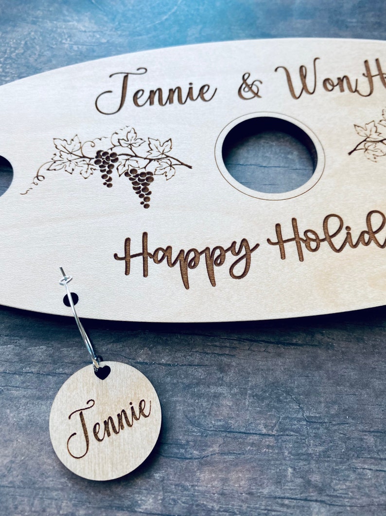 May include: Personalized wooden serving tray with two wine glass holders and a grapevine design. The tray says "Jennie & Won" and "Happy Holidays". A small wooden tag with the name "Jennie" is attached to the tray.