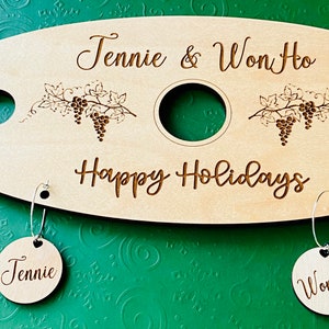 May include: A wooden wine carrier with two wine glass holders and a bottle holder. The carrier is engraved with the text "Jennie & Won Ho" and "Happy Holidays". Two wooden tags with the names "Jennie" and "Won Ho" are attached to the carrier.