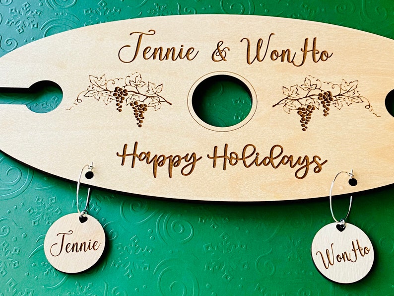 May include: A wooden wine carrier with two wine glass holders and a bottle holder. The carrier is engraved with the text "Jennie & Won Ho" and "Happy Holidays". Two wooden tags with the names "Jennie" and "Won Ho" are attached to the carrier.