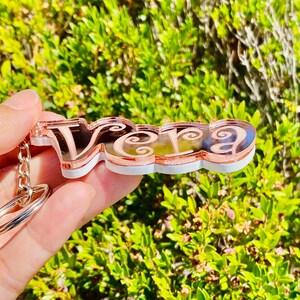 May include: A rose gold keychain with the name "Vera" in a decorative script font. The keychain is made of acrylic and has a white backing.