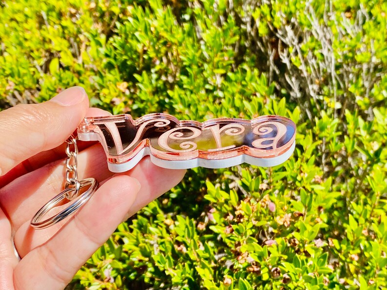 May include: A rose gold keychain with the name "Vera" in a decorative script font. The keychain is made of acrylic and has a white backing.
