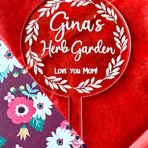 Puede incluir: Marcador de jardín de acrílico blanco con un diseño de corona y el texto "Gina's Herb Garden Love You Mom!"
