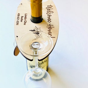 May include: Wooden wine bottle and glass holder with the text "Welcome Home!" and "Linda Chu Realtor 408-621-3296" engraved on it. The holder has a cutout for a bottle and two slots for wine glasses.