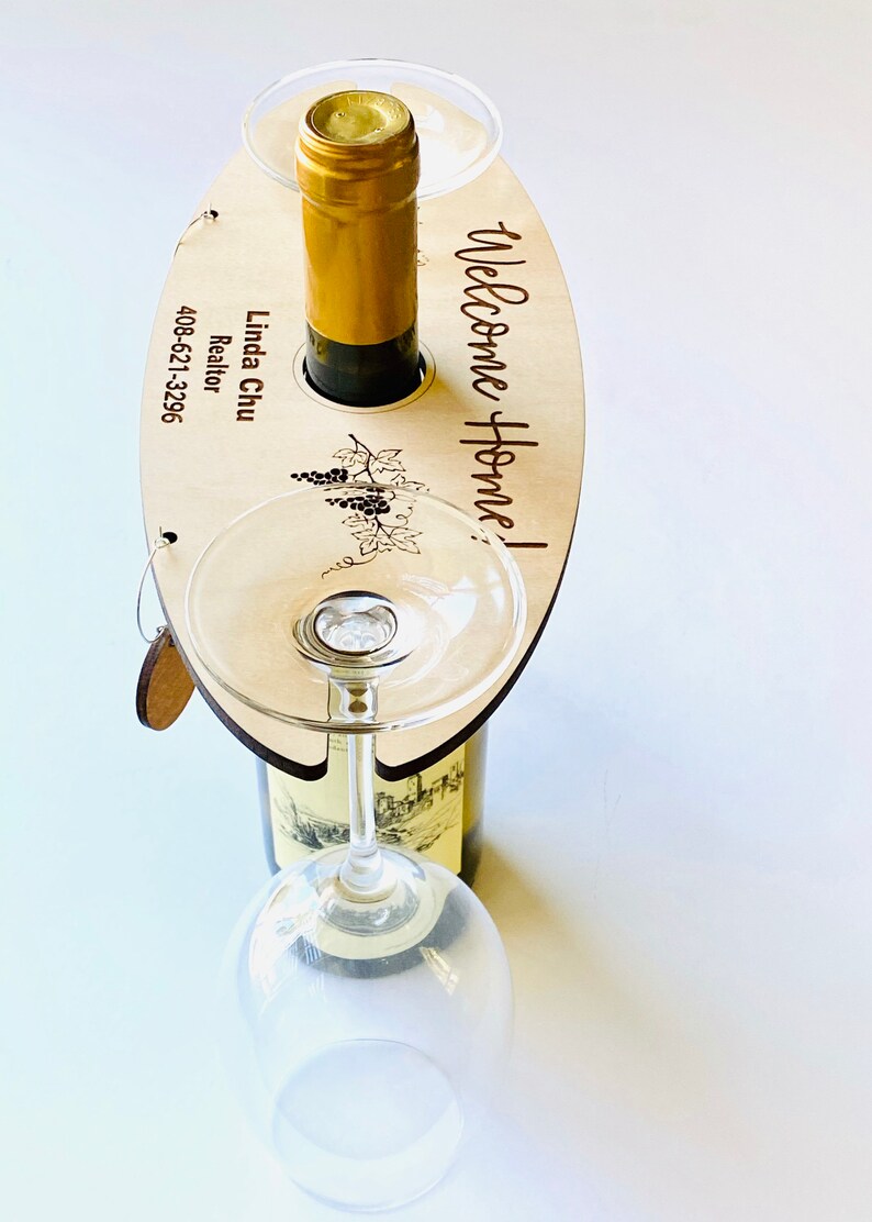 May include: Wooden wine bottle and glass holder with the text "Welcome Home!" and "Linda Chu Realtor 408-621-3296" engraved on it. The holder has a cutout for a bottle and two slots for wine glasses.