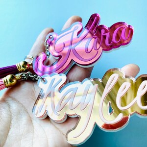May include: Two acrylic keychains with names "Kara" and "Kaylee" written in a cursive font. The keychain with the name "Kara" is pink and the keychain with the name "Kaylee" is gold. Both keychains have a pink and purple tassel.
