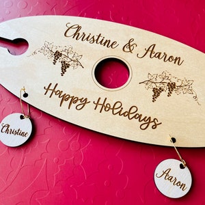 May include: Personalized wooden wine bottle holder with the text "Christine & Aaron Happy Holidays". Two round wooden tags with the names "Christine" and "Aaron" are attached.