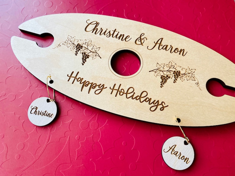 May include: Personalized wooden wine bottle holder with the text "Christine & Aaron Happy Holidays". Two round wooden tags with the names "Christine" and "Aaron" are attached.