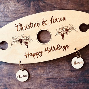 May include: Personalized wooden wine bottle holder with two hanging tags. The holder features a grapevine design and the text "Christine & Aaron Happy Holidays". The tags are labeled "Christine" and "Aaron".