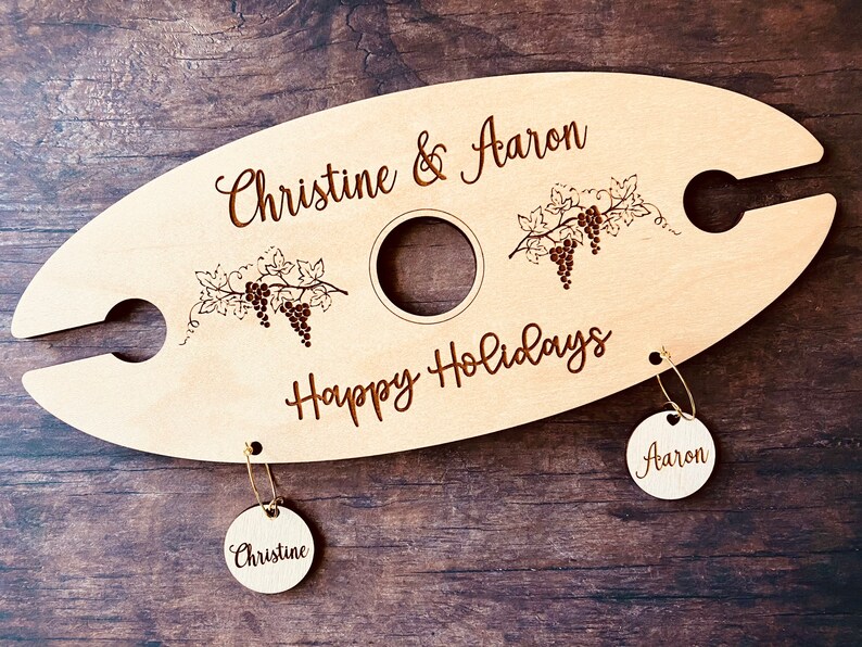May include: Personalized wooden wine bottle holder with two hanging tags. The holder features a grapevine design and the text "Christine & Aaron Happy Holidays". The tags are labeled "Christine" and "Aaron".