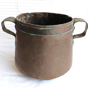 Puede incluir: Una olla de metal color cobre con dos asas curvas. La olla tiene un cuerpo ancho y redondeado y un borde ligeramente ensanchado. El metal muestra signos de edad y desgaste, con algunas zonas de oxidación verde.