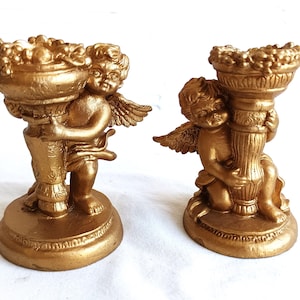 Puede incluir: Dos figuras de querubines dorados, cada una sosteniendo un cuenco decorativo. Los querubines tienen alas y están arrodillados. Los cuencos parecen estar llenos de fruta o flores. Las figuras están sobre una base redonda.