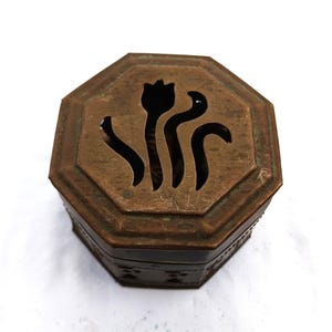 Puede incluir: Una caja octogonal de bronce antiguo con un diseño floral recortado en la tapa. La caja tiene una pátina oscura y envejecida, y los lados presentan un patrón repetido. La parte superior tiene una flor estilizada y líneas curvas.