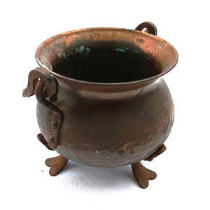 Puede incluir: Un pequeño caldero de cobre antiguo con una textura martillada. El caldero tiene dos asas y tres patas pequeñas. El cobre tiene una pátina oscura y envejecida con toques verdes. El caldero probablemente se utiliza con fines decorativos.