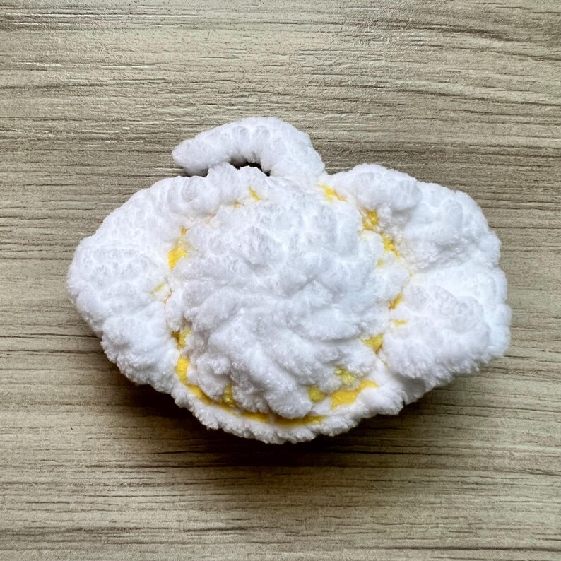 May include: A handmade, white, cloud-shaped crochet item with yellow accents. The fluffy, textured piece has a loop on top, suggesting it may be a decorative item or a toy. The item is placed on a wooden surface.