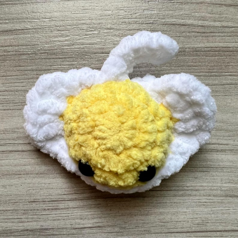 May include: A handmade, plush, crocheted egg-shaped toy with a yellow yolk and white surrounding the yolk. The toy has two black button eyes. The toy is on a wooden surface.