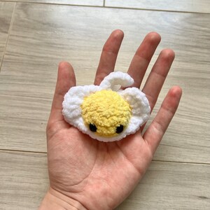 May include: A handmade crochet egg with a yellow yolk and white surrounding. The egg has two small black eyes. The egg is a cute, whimsical design.