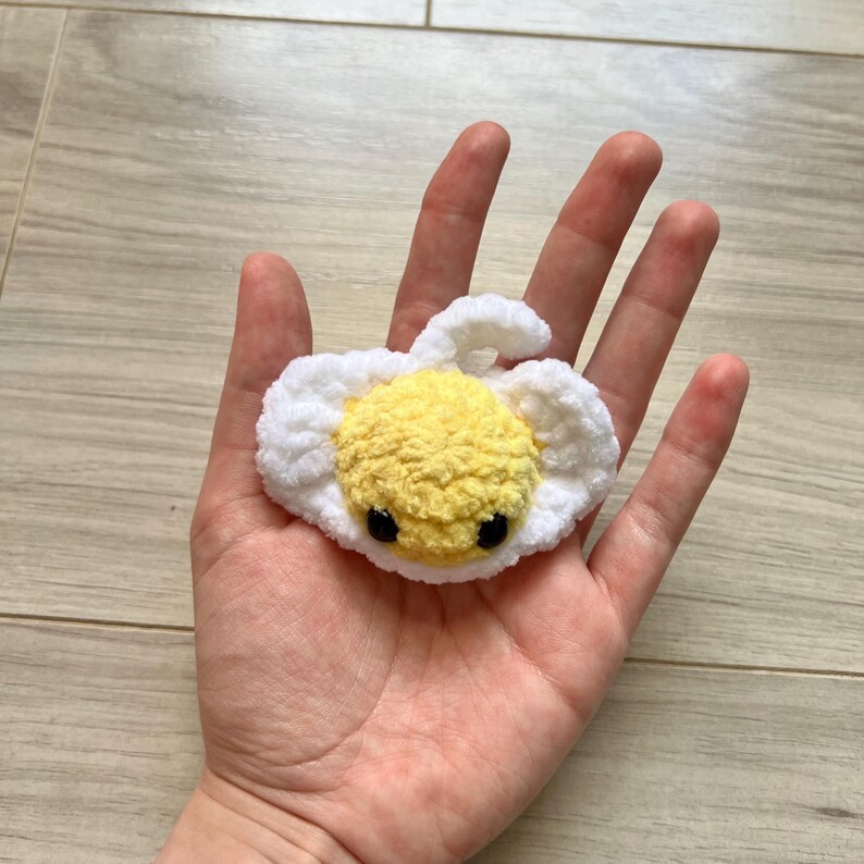 May include: A handmade crochet egg with a yellow yolk and white surrounding. The egg has two small black eyes. The egg is a cute, whimsical design.