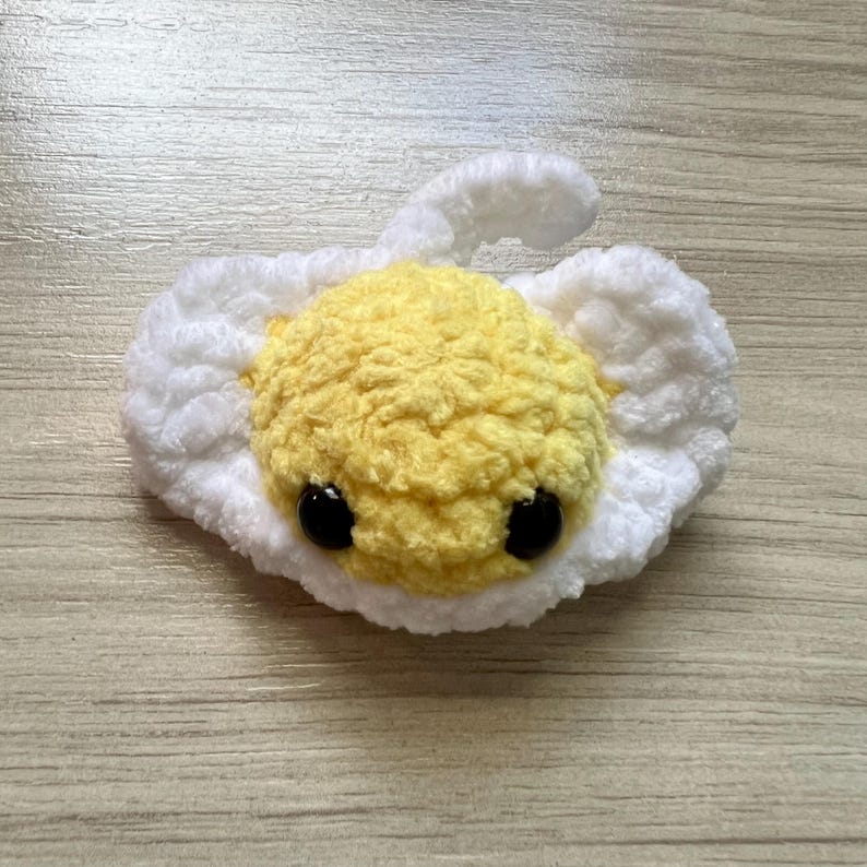 May include: A handmade, plush fried egg with a yellow yolk and white egg whites. The yolk has two black bead eyes. The egg is made of soft, textured yarn and is designed to look like a cartoon character.