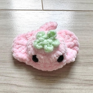 May include: A handmade pink crochet stingray with a green flower accent on top. The stingray has two black button eyes and a curled tail. The plush toy is made of soft, textured yarn.