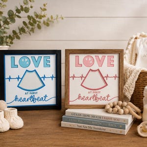 May include: Two framed signs with the words "LOVE" and "AT FIRST heartbeat" in blue and pink. A baby onesie, bottle, pacifier, and books are also present. A stuffed bunny and baby booties are on the left.