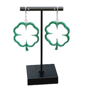 May include: A pair of green glitter four-leaf clover earrings on a black stand.