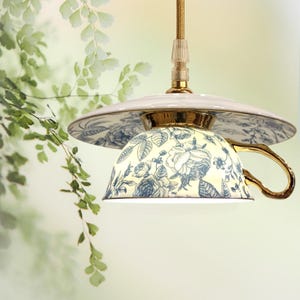 Könnte beinhalten: Eine einzigartige Pendelleuchte aus einer Teetasse und Untertasse. Die Tasse hat ein blaues Blumenmuster auf cremefarbenem Grund, mit goldenem Henkel und Zierleisten. Die Untertasse und die Beschläge sind ebenfalls goldfarben.