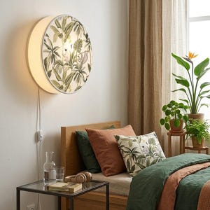 Puede incluir: Una lámpara de pared redonda con un diseño de jungla, con hojas de palmera verdes y blancas y un perezoso. La lámpara está montada en una pared blanca sobre una cama de madera con ropa de cama y almohadas verdes y óxido. Una mesita de noche con tapa de cristal y una jarra de agua está al lado de la cama.