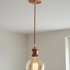 Puede incluir: Una lámpara colgante de color cobre con una bombilla grande y redonda de estilo Edison. La bombilla está encerrada en un accesorio de color cobre y cuelga de un cable trenzado marrón. El accesorio está montado en una placa de techo de color cobre.