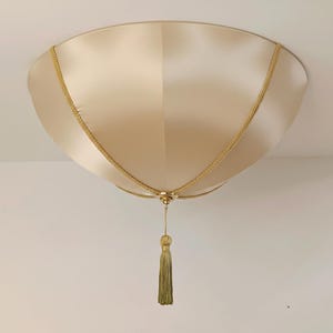 Pode incluir: Um candeeiro de teto de montagem semi-embutida com um abajur de tecido acetinado na cor champanhe. O abajur é acentuado com um acabamento trançado dourado decorativo que converge num ponto central, finalizado com uma borla dourada a combinar.