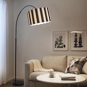 Puede incluir: Una lámpara de pie de arco negro con una pantalla a rayas. La pantalla tiene rayas verticales en negro, blanco y beige. La lámpara está en un salón, con un sofá y obras de arte en el fondo.