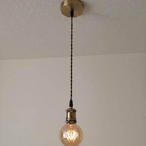 May include: A brass pendant light with a round, amber bulb. The light hangs from a brown, twisted cord and a matching brass ceiling mount. The bulb features a decorative filament. The light is suitable for a room.